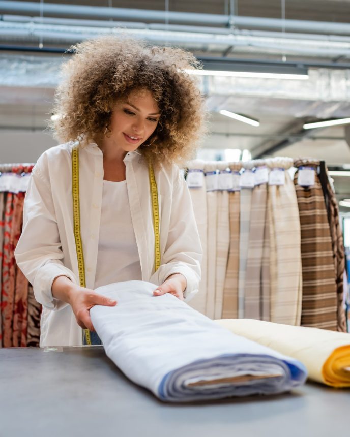 smiling saleswoman with measuring tape holding blue linen fabric roll in textile shop